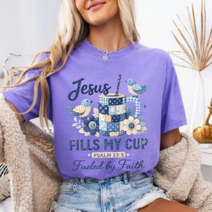 May include: A light purple t-shirt featuring a graphic of a blue patchwork mug, two small birds, and flowers. The text reads: Jesus FILLS MY CUP, PSALM 23:5, and Fueled by Faith. The design uses shades of blue, white, and yellow on the fabric.
