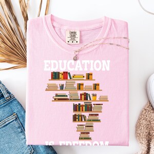 May include: A light pink t-shirt with the word "EDUCATION" in white block letters. Below the word is a graphic of bookshelves filled with colorful books. The phrase "IS FREEDOM" is at the bottom of the shirt.