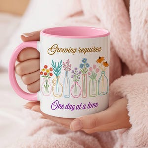May include: White ceramic mug with a pink interior and handle. The mug features the text "Growing requires" and "One day at a time" with colorful illustrations of flowers in vases. The mug is held by a person wearing a pink robe.