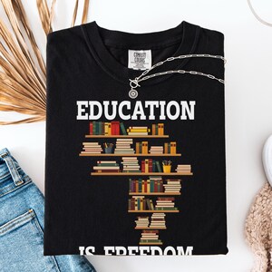 May include: Black t-shirt with the word "EDUCATION" in white block letters. Below the word is a graphic of bookshelves filled with colorful books. The phrase "IS FREEDOM" is printed in white block letters at the bottom of the shirt.
