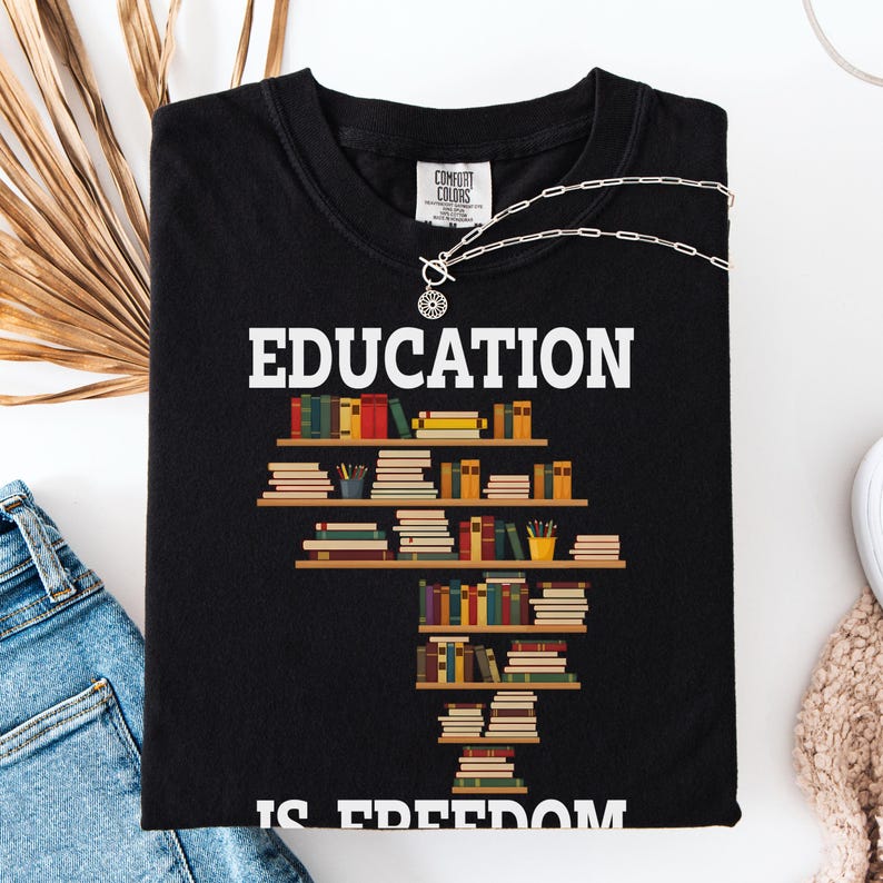 May include: Black t-shirt with the word "EDUCATION" in white block letters. Below the word is a graphic of bookshelves filled with colorful books. The phrase "IS FREEDOM" is printed in white block letters at the bottom of the shirt.