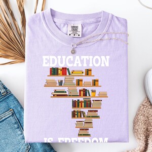 May include: A light purple t-shirt with the word "EDUCATION" in white block letters. Below the word is a graphic of a bookshelf filled with books. The phrase "IS FREEDOM" is printed below the bookshelf graphic.