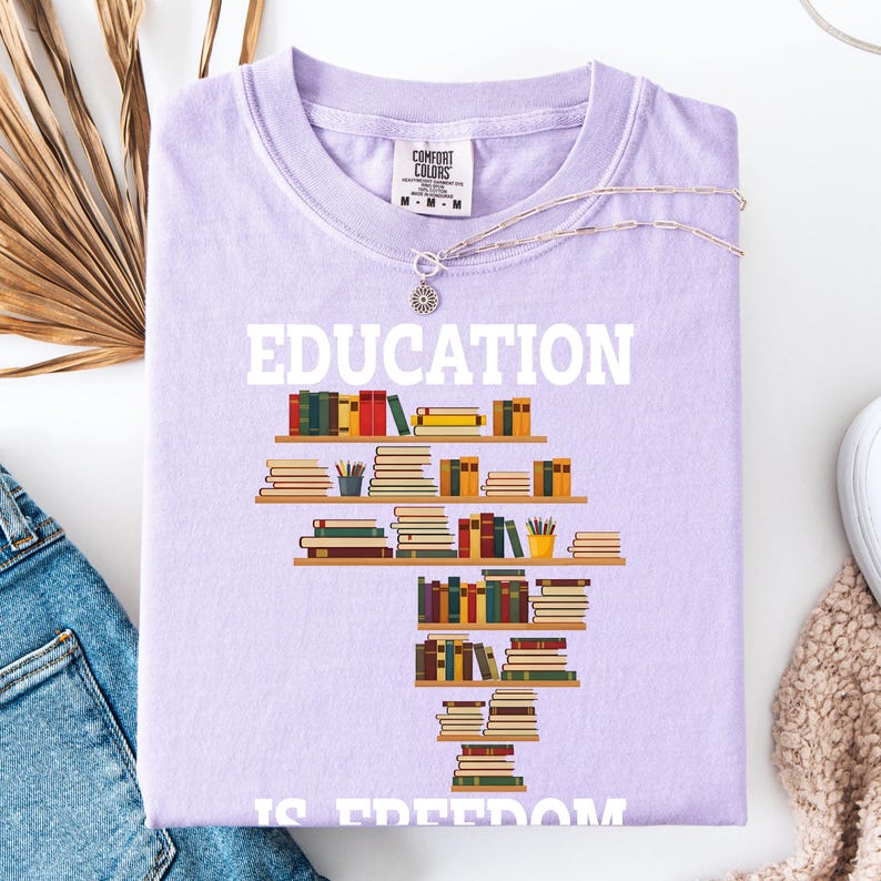 May include: A light purple t-shirt with the word "EDUCATION" in white block letters. Below the word is a graphic of a bookshelf filled with books. The phrase "IS FREEDOM" is printed below the bookshelf graphic.