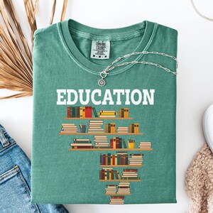May include: A teal Comfort Colors t-shirt with the word "EDUCATION" in white block letters. Below the word is a graphic of bookshelves filled with books. A silver necklace with a sunburst pendant is on the shirt.
