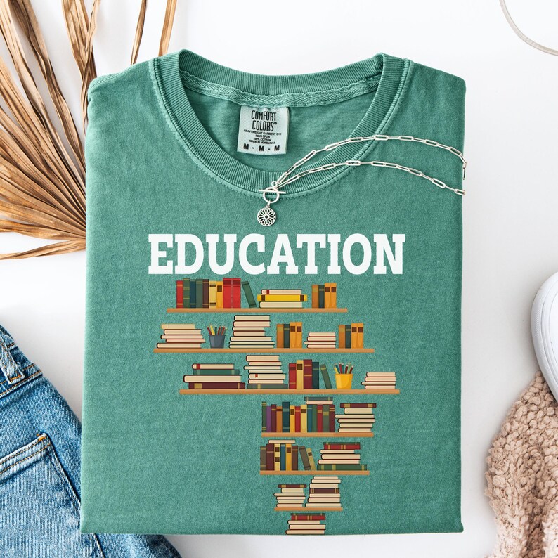May include: A teal Comfort Colors t-shirt with the word "EDUCATION" in white block letters. Below the word is a graphic of bookshelves filled with books. A silver necklace with a sunburst pendant is on the shirt.