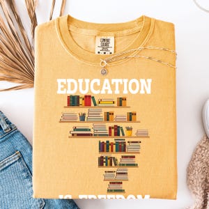 May include: Mustard yellow t-shirt with the word "EDUCATION" in white block letters. A graphic of bookshelves filled with books is below the word. A silver necklace with a sunburst pendant is draped across the shirt.