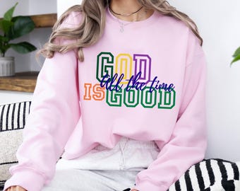 May include: A light pink crewneck sweatshirt featuring the text GOD IS GOOD in block letters with green, yellow, orange, and purple colors, and the phrase all the time written in a blue cursive script across the center of the design.