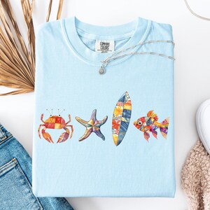 May include: Light blue Comfort Colors t-shirt featuring a watercolor design of a crab, starfish, surfboard, and fish. The design incorporates a patchwork pattern in various colors. A silver necklace with a sun pendant is draped across the shirt.