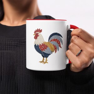 May include: White ceramic mug with a red interior and handle. The mug features a colourful rooster design with patchwork-style details in red, blue, and yellow. The rooster has a red comb and wattle.