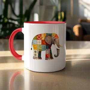 Puede incluir: Taza de cerámica blanca con interior y asa rojos. La taza presenta un colorido diseño de elefante patchwork. El elefante tiene una cabeza y colmillos marrones, y su cuerpo está formado por varios patrones geométricos multicolores.