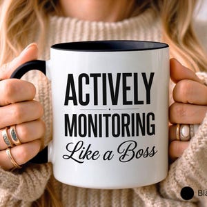 May include: White ceramic mug with a black interior and handle. The mug features the text "ACTIVELY MONITORING Like a Boss" in black, bold font. The mug is held by a person wearing rings.