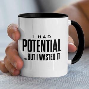 May include: A white ceramic mug with a black handle and the text "But First, Coffee" in a black, handwritten-style font. The mug is on a white surface.