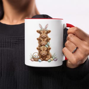May include: White ceramic mug with a red interior and handle. The mug features a stack of three brown Highland cows, decorated with Easter bunny ears, flowers, and bows. A basket of Easter eggs and colorful eggs are also shown.