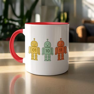 May include: White ceramic mug with a red interior and handle. The mug features three colourful robot illustrations in yellow, green, and orange, each with a number. The robots are numbered 01, 10, and 11.