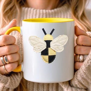 以下が含まれることがあります： 黄色い内側とハンドルが付いた白いセラミックマグ。マグには、黄色のギンガムと黒のパッチ、クリーム色の羽が付いた布製の蜂のアップリケが付いています。蜂のデザインはマグの前面の中央に配置されています。
