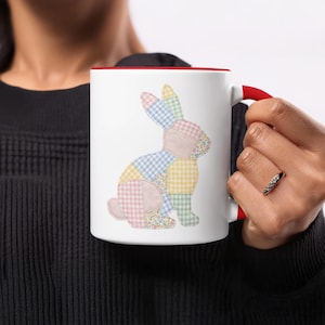 Puede incluir: Taza de cerámica blanca con interior y asa rojos, con un diseño de conejo de retazos. El conejo está compuesto por varios patrones de tela de colores pastel, incluyendo cuadros y estampados florales. Un diseño encantador y caprichoso.