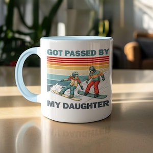 May include: White ceramic mug with a light blue handle and interior. The mug features a retro design with the text "GOT PASSED BY MY DAUGHTER" above an illustration of two snowboarders on a mountain slope.