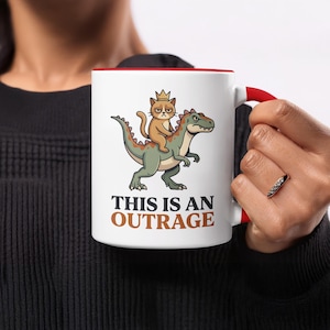 May include: White ceramic mug with a red interior and handle. The mug features a cartoon illustration of a cat wearing a crown riding a dinosaur. The text "THIS IS AN OUTRAGE" is printed below the illustration.