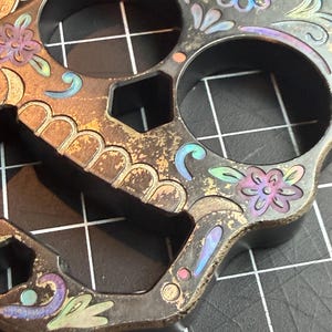 May include: A dark metal skull-shaped tool featuring two large circular finger holes and engraved floral patterns with iridescent blue, purple, and gold accents. The surface has a weathered, distressed finish with visible metallic texture.