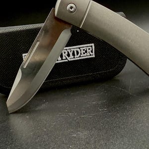 May include: A folding pocket knife with a metallic silver handle and a partially extended steel blade. The knife is positioned in front of a black fabric carrying case that features the white embroidered text RYDER on its front surface.