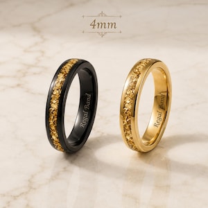 Könnte beinhalten: Zwei 4 mm Ringe, einer in Schwarz und einer in Gold, jeweils mit einer zentralen Einlage aus zerkleinerten goldfarbenen Steinfragmenten. Beide Ringe sind innen mit dem Text Regal Band graviert und haben eine polierte Oberfläche.