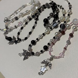 May include: Several beaded necklaces with angel wing and star charms. The necklaces feature a variety of beads in colors like white, black, pink, and gray. The chains are silver-toned, and the necklaces are displayed on a white surface.