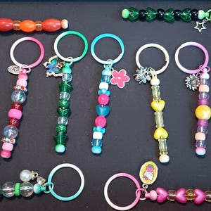 May include: A collection of beaded keychains in various colors, including pink, green, blue, and yellow. Some keychains feature heart, star, and butterfly charms. The keychains have metal rings and are arranged on a dark surface.
