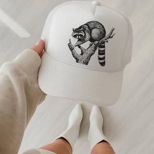 Puede incluir: Gorra de camionero blanca con una ilustraci&oacute;n en blanco y negro de un mapache posado en una rama de &aacute;rbol. La gorra tiene una parte trasera de malla y una visera curva. El dise&ntilde;o del mapache es detallado y est&aacute; centrado.