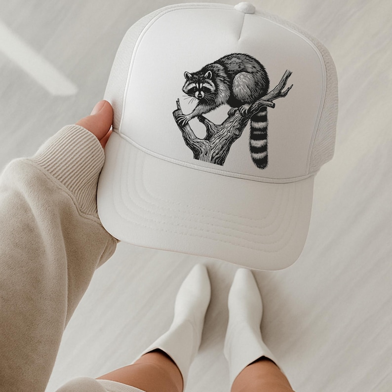 Puede incluir: Gorra de camionero blanca con una ilustraci&oacute;n en blanco y negro de un mapache posado en una rama de &aacute;rbol. La gorra tiene una parte trasera de malla y una visera curva. El dise&ntilde;o del mapache es detallado y est&aacute; centrado.