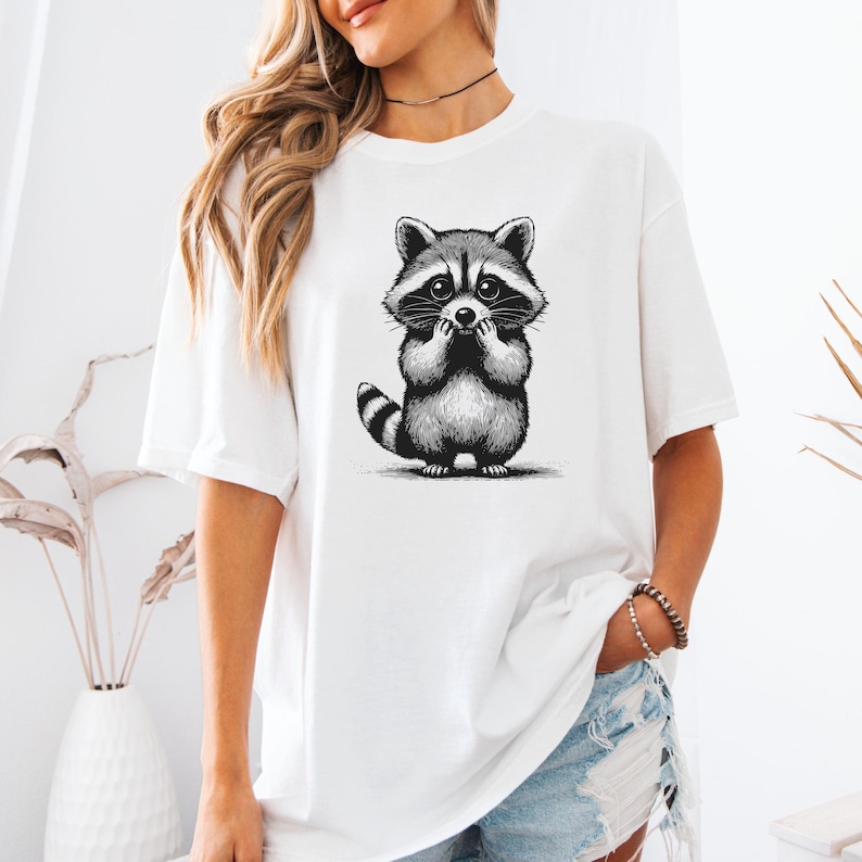 Puede incluir: Camiseta blanca con una ilustraci&oacute;n en blanco y negro de un mapache. El mapache est&aacute; representado con las manos cerca de la boca, mostrando una expresi&oacute;n de sorpresa o timidez. La camiseta es de estilo casual.