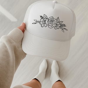 Pu&ograve; includere: Cappellino da camionista bianco con design floreale. Il cappellino ha un pannello frontale bianco e un retro in rete. Un design floreale nero, tra cui rose e foglie, &egrave; centrato sulla parte anteriore.