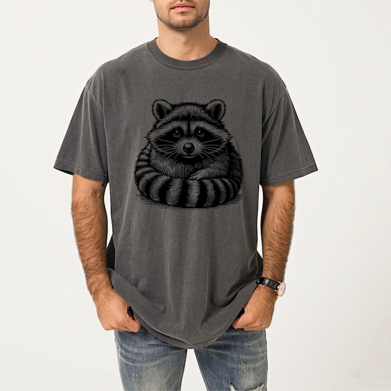 Puede incluir: Camiseta gris oscuro con una ilustraci&oacute;n en blanco y negro de un mapache. El mapache est&aacute; representado sentado con las patas cruzadas. La camiseta tiene mangas cortas y un corte holgado.