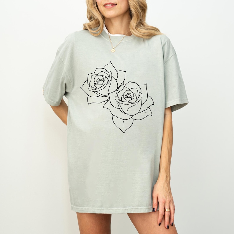 Pu&ograve; includere: T-shirt verde chiaro con un contorno nero di due rose. Le rose sono dettagliate con petali e foglie. La maglietta ha maniche corte e una vestibilit&agrave; ampia.
