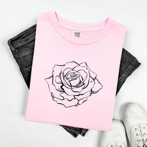 Pu&ograve; includere: Una maglietta rosa chiaro con una grande rosa nera e bianca. La maglietta &egrave; appoggiata su jeans neri. Le scarpe da ginnastica bianche sono visibili in basso. L'etichetta sulla maglietta dice "Comfort Colors".