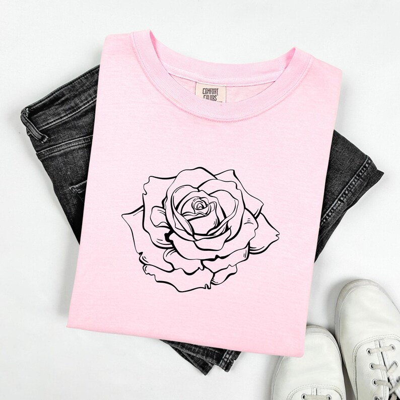 Pu&ograve; includere: Una maglietta rosa chiaro con una grande rosa nera e bianca. La maglietta &egrave; appoggiata su jeans neri. Le scarpe da ginnastica bianche sono visibili in basso. L'etichetta sulla maglietta dice "Comfort Colors".