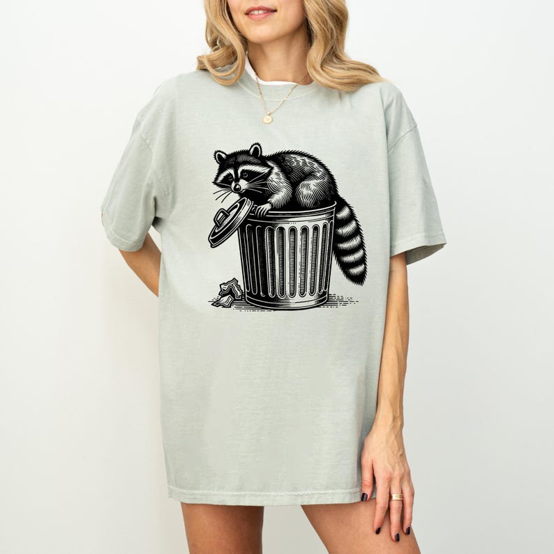 Puede incluir: Camiseta verde claro con una ilustraci&oacute;n en blanco y negro de un mapache sentado en un cubo de basura. El mapache tiene una cola rayada y est&aacute; mirando hacia afuera. El cubo de basura tiene una tapa y est&aacute; rodeado de basura.