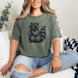 Puede incluir: Camiseta verde oliva con una ilustraci&oacute;n detallada de un mapache y una nota musical. El mapache est&aacute; representado con las patas juntas, pareciendo cantar. La camiseta se combina con unos vaqueros azules rotos.