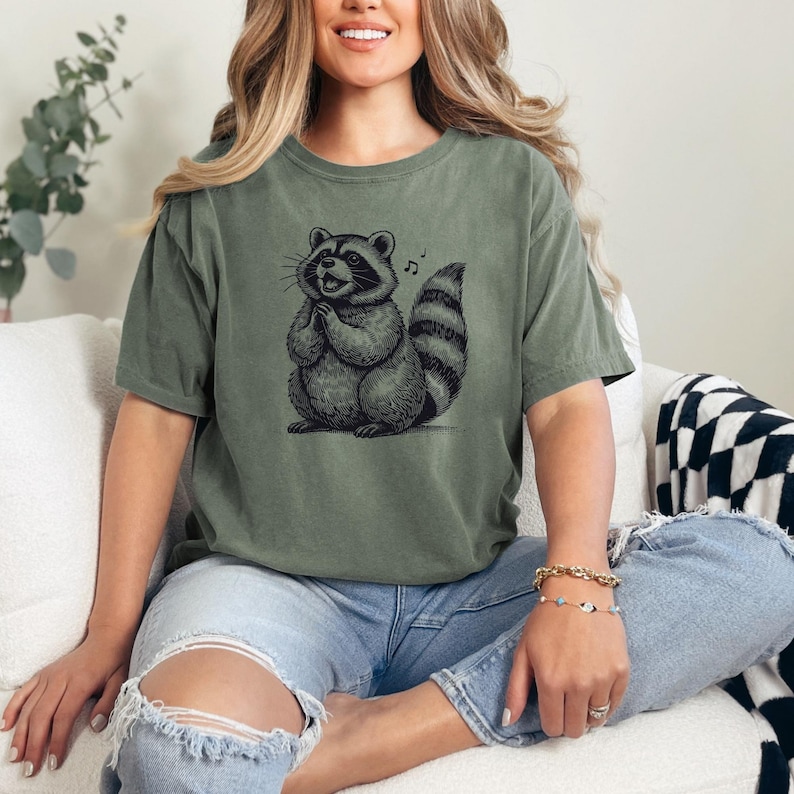 Puede incluir: Camiseta verde oliva con una ilustraci&oacute;n detallada de un mapache y una nota musical. El mapache est&aacute; representado con las patas juntas, pareciendo cantar. La camiseta se combina con unos vaqueros azules rotos.
