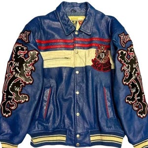 May include: Blue leather jacket with red and white stripes, featuring embroidered black lion graphics on the sleeves. The jacket has a button-up front, a collar, and a cream-colored stripe across the chest. The jacket has the text "FLY HIGH" and "BLOOD WOLF".