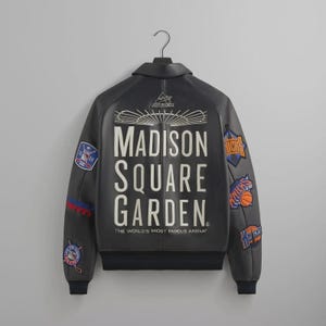 May include: A black leather jacket with the text "MADISON SQUARE GARDEN" in white. The jacket has multiple patches with team logos and the words "THE WORLD'S MOST FAMOUS ARENA" at the bottom.
