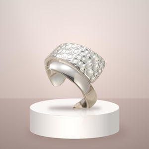 May include: A silver-toned ring featuring a wide band with a textured, hammered surface pattern on the upper portion and a smooth, polished finish on the lower section. The design consists of two distinct segments that overlap to form the ring.