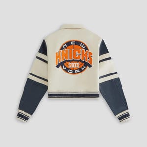 May include: Cream-colored varsity jacket with navy blue sleeves and striped trim. The back features a circular logo with the text "New York Knicks" in orange and navy blue, with the brand name "Kith" below.