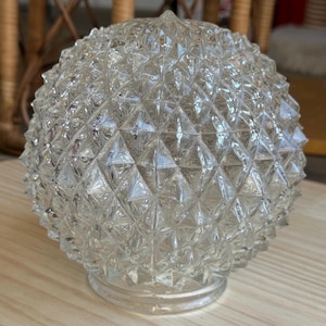 May include: Clear glass globe with a diamond-cut pattern. The round light fixture has a textured surface and a circular base. The glass is transparent, allowing light to pass through. This decorative piece is suitable for home decor.