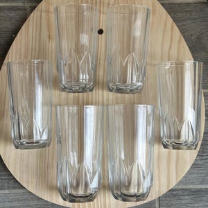 May include: Set of six clear, patterned drinking glasses. The glasses are arranged on a light-colored wooden surface. Each glass has a vertical ribbed design and a decorative pattern near the base.