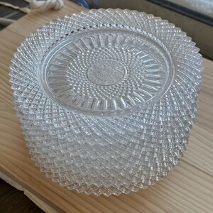 May include: A stack of clear, pressed glass plates with a diamond pattern and a central floral design. The plates are arranged on a light-colored wooden surface, showcasing their intricate details and reflective qualities.