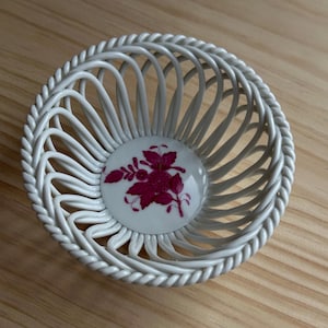 May include: A white, openwork porcelain basket with a floral design. The basket features a woven design with a decorative, twisted rim. The center of the basket has a white background with a magenta floral pattern.