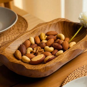 Può includere: Una ciotola di legno piena di una varietà di noci, tra cui mandorle, anacardi e noci. La ciotola è posta su un tavolo di legno, con un fiore bianco. Sono visibili anche un piatto e un sottobicchiere intrecciato.