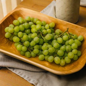 May include: A wooden serving bowl filled with green grapes. The bowl is rectangular with rounded corners and a natural wood grain finish. The grapes are fresh and ripe, with a vibrant green colour.