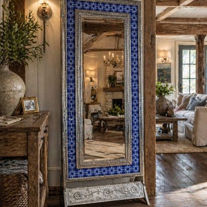 May include: A tall, rectangular mirror with a silver frame and a blue and white patterned border. The mirror is standing on a decorative metal base with floral accents. The mirror reflects a room with a fireplace and furniture.