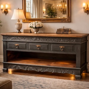 May include: A dark gray and wood-toned console table with three drawers and ornate carvings. The table has brass ring pulls and a lower shelf. A lamp, flowers, and books are on top, with a mirror in the background. The room has sconces and a neutral color palette.